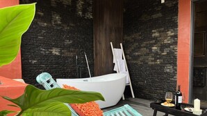 Un espacio de spa con una bañera grande, un banco de madera con cojín naranja, y una mesita con vela y copas de vino.