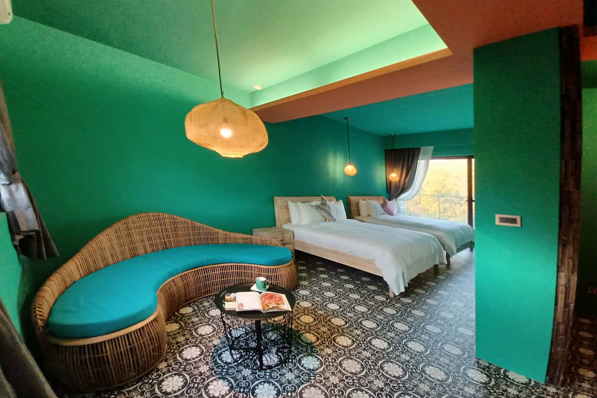 Ein Zimmer mit türkisfarbenen Wänden, einem Bett mit weißer Bettwäsche, einem Korbssessel mit türkisfarbenen Kissen, einem gemusterten Boden und einer hängenden Leuchte.