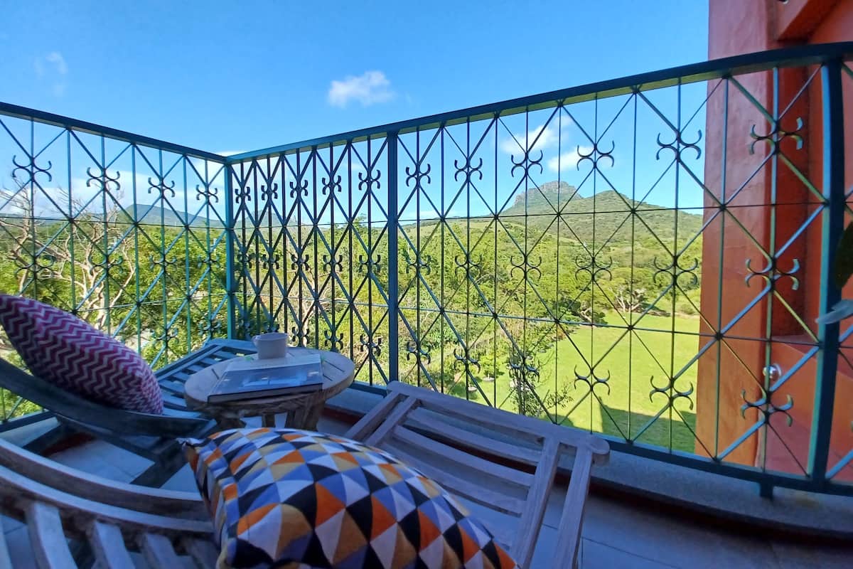 Ein Balkon mit Blick auf eine grüne Landschaft und Berge. Dort steht ein Tisch mit einem Stuhl und einem gemusterten Kissen.