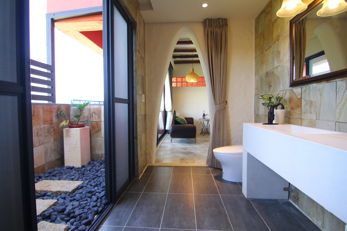 Ein modernes Badezimmer mit einem großen Waschbecken, Toilette und einer Duschkabine. Auf der Ablage steht ein Blumentopf und durch eine geöffnete Tür ist der Wohnbereich sichtbar.