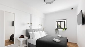 A modern bedroom with a large bed, a bedside table, a mirror, a small window with plants, and a TV mounted on the wall. - A Modern and Homely 2BD Apartment in TLV (Tel Aviv)
