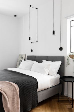 Ein modernes Schlafzimmer mit grauem Bett, weißen Kissen, einem schwarzen Nachttisch und einer hängenden Leuchte.