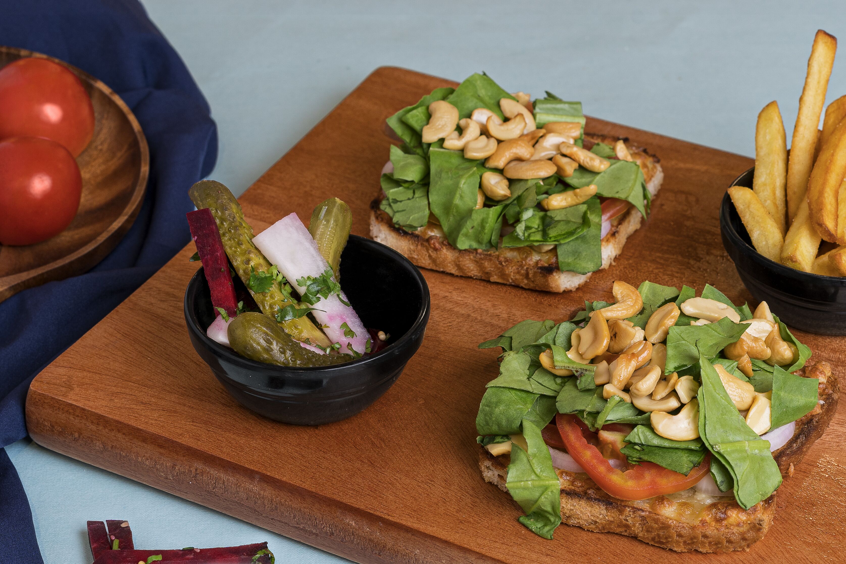 Twee belegde sandwiches op een houten plank, samen met een kom augurken en een kom frieten.