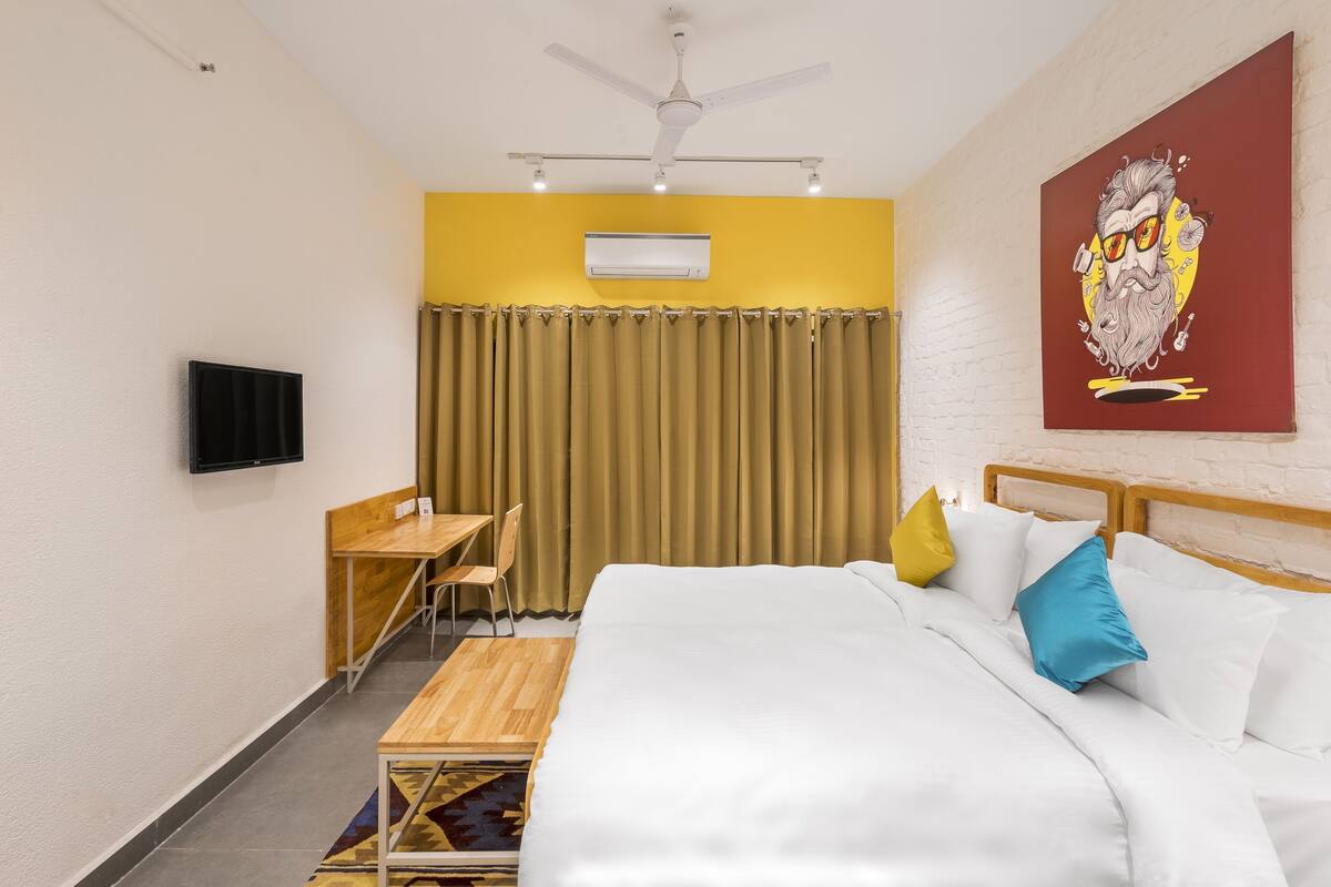 A hotel room with a bed, a desk, a chair, a ceiling fan, an air conditioner, and a TV.