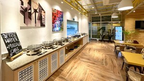 A modern cafe with a long counter displaying various food and drink options, a chalkboard menu, and a large window offering a view of the outside. - Bedzzz Varanasi by Leisure Hotels (Varanasi)