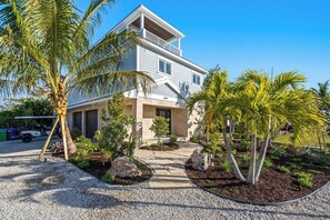 A two-story house with a balcony, surrounded by palm trees and a gravel driveway.