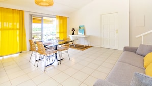 Un salon lumineux avec un coin repas, des rideaux jaunes, une porte blanche et un canapé agrémenté d’un coussin jaune.