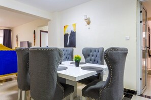 A dining area with a white table, four upholstered chairs with gold studs, a potted plant, and a wall-mounted light fixture. - Stay.Plus Mango Court Apartment Kilimani (Nairobi)