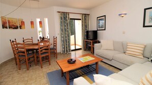 Uma sala de estar com uma mesa de jantar de madeira, cadeiras, um sofá, uma televisão e uma porta de vidro deslizante que dá para uma área externa.