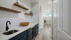A kitchen with white subway tiles, dark cabinets, a wooden shelf, and a basket on the counter. - Mango Bay – Luxury Island Home w/ Heated Pool, Spa & Walk To Beach (Holmes Beach)