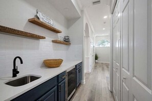 A kitchen with white subway tiles, dark cabinets, a wooden shelf, and a basket on the counter. - Mango Bay – Luxury Island Home w/ Heated Pool, Spa & Walk To Beach (Holmes Beach)