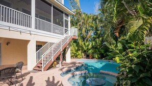 A two-story house with a balcony, a swimming pool with a hot tub, and lush tropical vegetation. - Mango Bay – Luxury Island Home w/ Heated Pool, Spa & Walk To Beach (Holmes Beach)