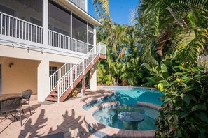 A two-story house with a balcony, a swimming pool with a hot tub, and lush tropical vegetation. - Mango Bay – Luxury Island Home w/ Heated Pool, Spa & Walk To Beach (Holmes Beach)