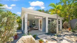 A single-story house with a covered porch, a patio with outdoor furniture, and a well-maintained garden with various plants and rocks. - The Cottage on Pine - Charming Pine Ave Cottage w/ Heated Pool in Heart of AMI (Anna Maria)