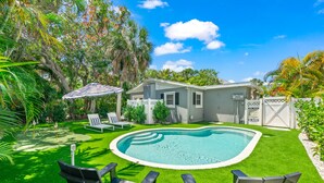 A backyard with a swimming pool, green artificial grass, and two lounge chairs. - The Cottage on Pine - Charming Pine Ave Cottage w/ Heated Pool in Heart of AMI (Anna Maria)