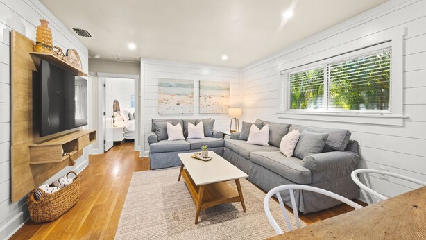 A modern living room with a sectional sofa, a flat-screen TV, and a wooden coffee table. - The Cottage on Pine - Charming Pine Ave Cottage w/ Heated Pool in Heart of AMI (Anna Maria)