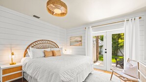 A bedroom with a large bed, white bedding, a wicker headboard, a bedside lamp, and a view of the outdoors through a sliding glass door. - The Cottage on Pine - Charming Pine Ave Cottage w/ Heated Pool in Heart of AMI (Anna Maria)