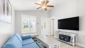 A living room with a blue sofa, a coffee table, a TV on a white cabinet, and a ceiling fan. - Point Three + Pool, Bikes, Nearby Beach & Sunroom (Panama City Beach)