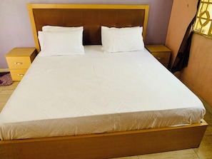 Un llit ben fet amb capçalera de fusta en una habitació amb una paret morada i una petita taula de nit de fusta a prop.