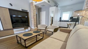 Un salon moderne avec un téléviseur à écran plat, une table basse en bois, des canapés de couleur beige et un coin cuisine à l’arrière-plan.