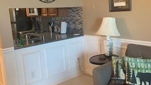 Een woonkamer met een bank, een klein rond tafeltje, een lamp, een decoratief wandobject, en een keuken met een spoelbak en kastjes.