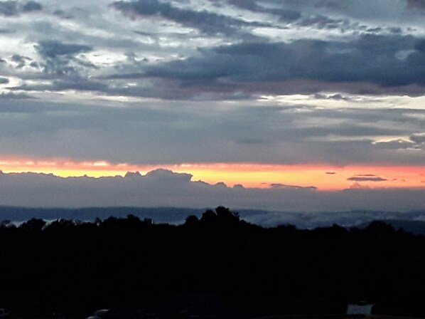 Un paysage avec un coucher de soleil sur une chaîne de montagnes et la silhouette d’arbres au premier plan.
