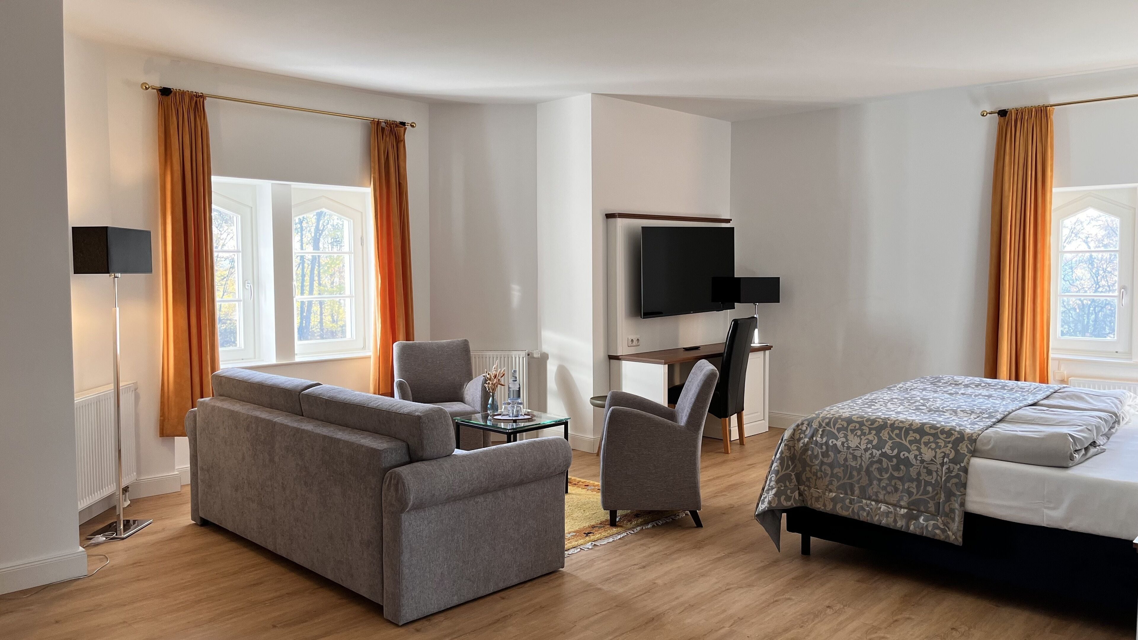 A hotel room with a sofa, a desk with a chair, a bed, a TV, and large windows with curtains.