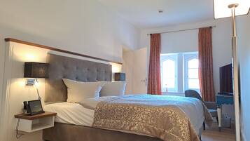 A hotel room with a large bed, bedside table with a lamp, a desk with a chair, and a window with curtains.