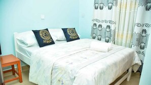 A neatly made bed with white bedding and two pillows featuring a gold emblem, a wooden nightstand, and a patterned curtain. - Stay.Plus Arujo Apartment Homa Bay (Homa Bay)