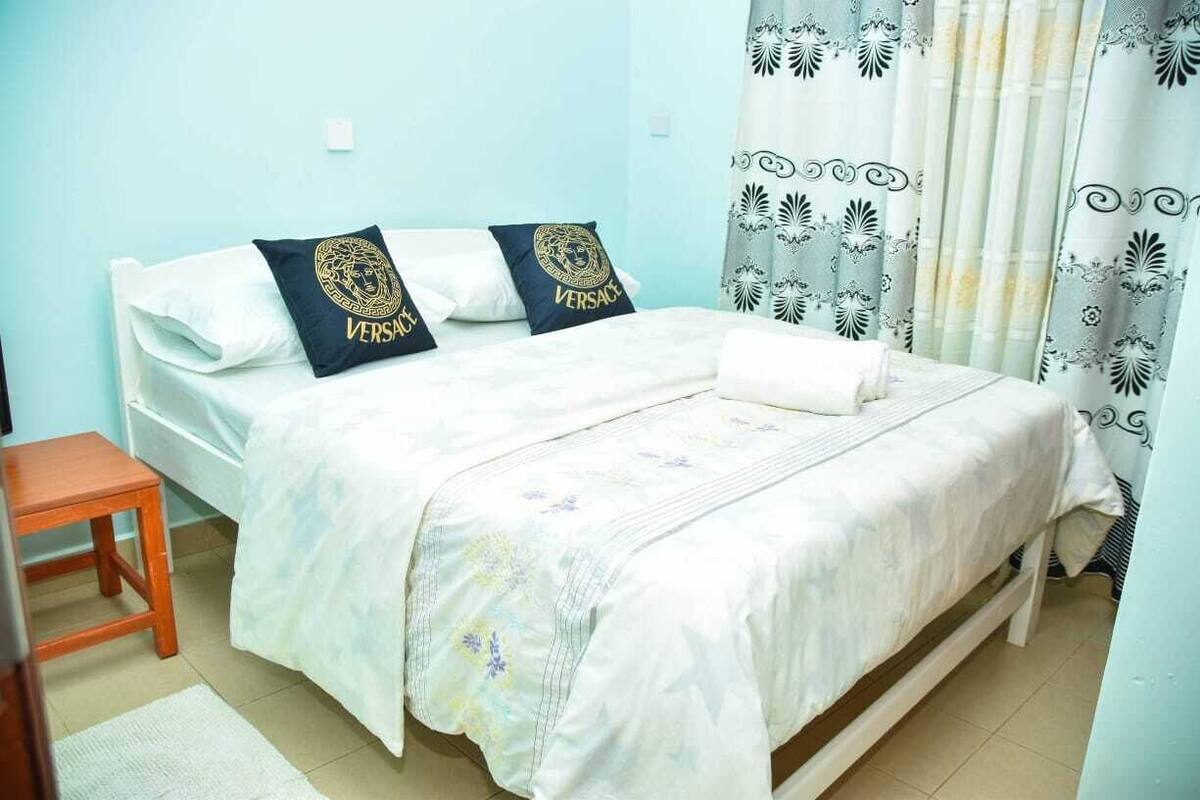 A neatly made bed with white bedding and two pillows featuring a gold emblem, a wooden nightstand, and a patterned curtain.