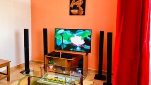 A living room with a flat-screen TV on a wooden stand, a glass coffee table with a floral centerpiece, and a red curtain to the right. - Stay.Plus Arujo Apartment Homa Bay (Homa Bay)