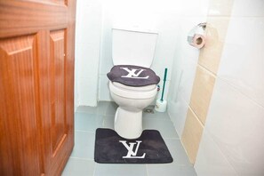 A bathroom with a toilet, a wooden door, a toilet paper holder, and a black mat with a white logo.