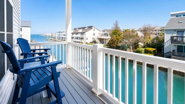 A wooden deck with blue chairs, a white railing, and a view of a marina with boats and houses. - Large Waterfront Townhome with Boat Dock and Pool (Ocean City)