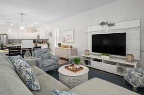 A modern living room with a large flat-screen TV, a sofa, a coffee table, and a kitchen in the background.