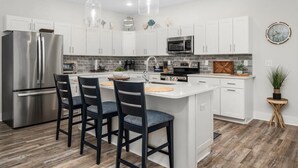 A modern kitchen with white cabinets, stainless steel appliances, a central island with bar stools, and a hanging pendant light. - Large Waterfront Townhome with Boat Dock and Pool (Ocean City)
