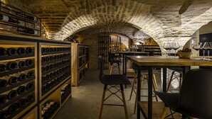 An underground wine cellar with stone arches, wooden shelves with wine bottles, and a table with glasses and a basket. - Hotel du Palais (Dijon)