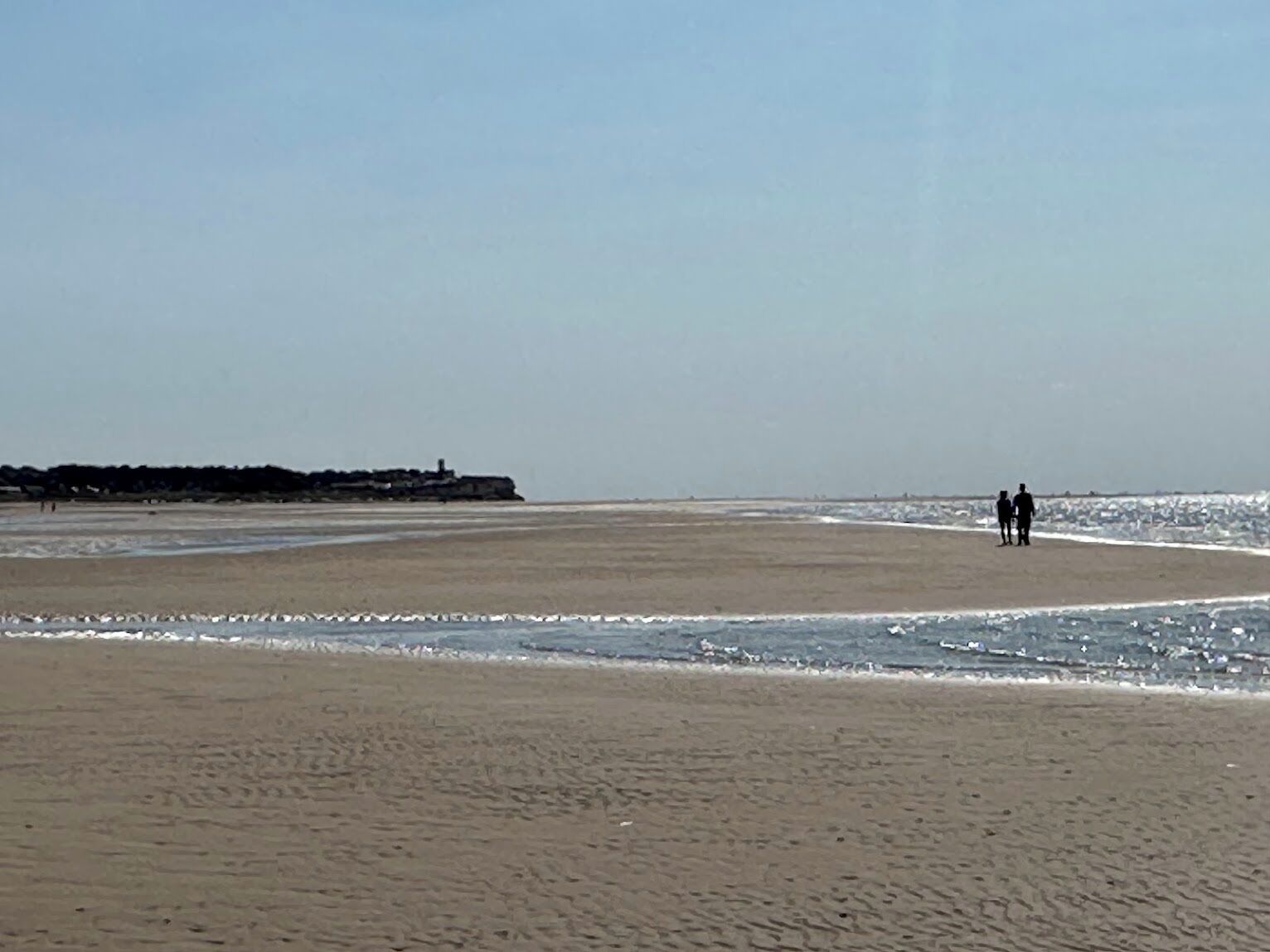 A wide beach with a rocky outcrop in the distance and two people walking along the shore.