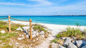Una playa con una cerca de madera y una barrera de cuerda, que conduce a una orilla arenosa con agua azul cristalina y una costa distante.