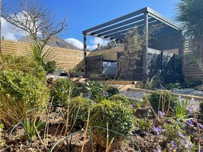 A modern outdoor patio area with a pergola, garden beds, and various plants.