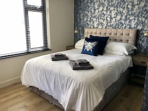 A neatly made bed with white linens and two folded towels, a blue pillow with a bee design, and a wooden nightstand with a lamp. - Captivating 5-bed House in Porthleven (Helston)
