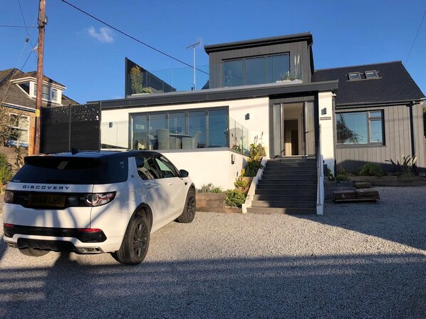 A modern two-story house with a white exterior and large windows. A white SUV is parked in front.