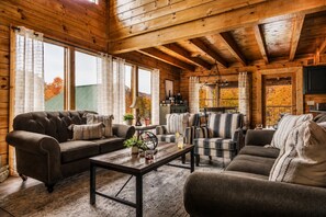 A cozy cabin interior with a wooden ceiling, large windows, a fireplace, and a television.
