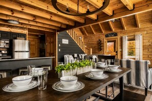 A wooden cabin interior with a dining area set for four, kitchen appliances, and a staircase leading to a loft.