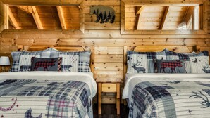 A cabin with two beds, each with plaid bedding and animal-themed pillows, wooden walls, and a sloped ceiling with a bear and trees cutout.
