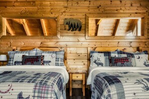 A cabin with two beds, each with plaid bedding and animal-themed pillows, wooden walls, and a sloped ceiling with a bear and trees cutout.
