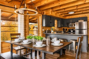 A wooden interior with a dining table set for four, kitchen area with appliances, and a view of autumn trees.