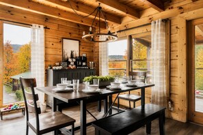 A wooden interior with a dining table set for four, a coffee station, and large windows offering a view of autumn foliage.