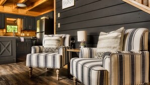 A cozy living room with striped armchairs and a small table, wooden beams on the ceiling, and a view of the kitchen through a window.