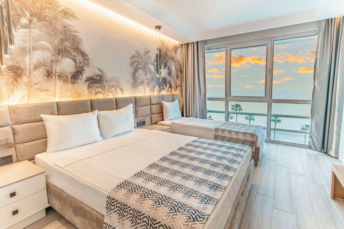 Une chambre d’hôtel avec deux lits, une grande fenêtre donnant sur le coucher de soleil et une fresque murale représentant des palmiers.