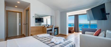A modern hotel room with a large bed, a desk, a TV, and a balcony with a view of the sea.
